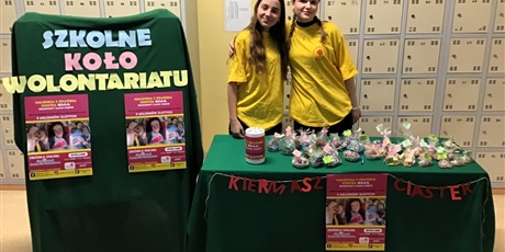 Powiększ grafikę: Dwie dziewczynki w żółtych bluzkach stoją przy stole, na stole leżą zapakowane w folię babeczki, po lewej stronie stoi tablica, na której jest napis Szkolne koło wolontariatu i dwa plakaty o akcji charytatywnej