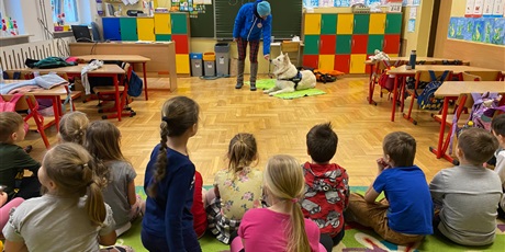 Powiększ grafikę: Grupa dzieci siedzących na dywanie a w tle stoi opiekun z psem ratownikiem Kajko