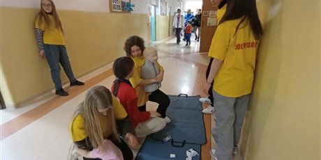 Powiększ grafikę: Na szkolnym korytarzu dwie uczennice w żółtych koszulkach klęczą ćwicząc odkrztuszanie dzieci na fantomach, jedna uczennica w czerwonej koszulce kuca przy nich, 3 uczniów się przyglada. W tle uczestnicy Dnia Otwartego.