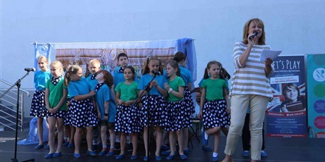 Powiększ grafikę: Na scenie kilkanaścioro śpiewających dzieci, z prawej strony z mikrofonem w dłoni Pani Dyrektor
