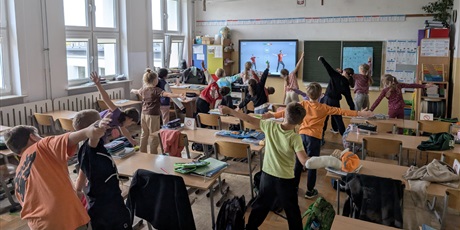 Powiększ grafikę: Uczniowie stoją obok swoich ławek i wykonują ćwiczenia ruchowe, naśladując postacie z filmu wyświetlanego na ekranie z przodu klasy.