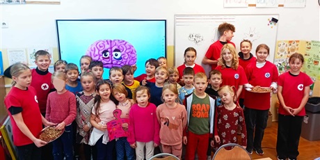 Powiększ grafikę: W sali lekcyjnej na tle tablicy multimedialnej z grafiką mózgu stoją ustawieni w trzy rzędy uczniowie klasy 1c i członkowie Koła PP HOPR. Pierwszy rząd uczniów trzyma plakat oraz tacę z orzechami.