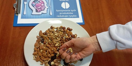 Powiększ grafikę: Na stole leży grafika dotycząca Udaru Mózgu. Na jej tle dłoń sięgająca po orzechy z talerza