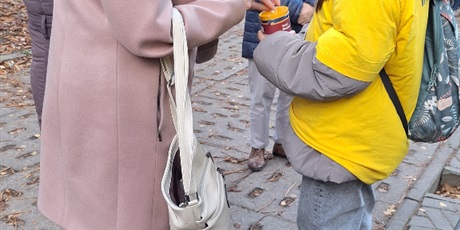 Powiększ grafikę: Trzy starsze kobiety stoją przy dziewczynce w koszulce wolontariusza, która w ręku trzyma skarbonkę. Jedna z nich wrzuca do puszki pieniądze.