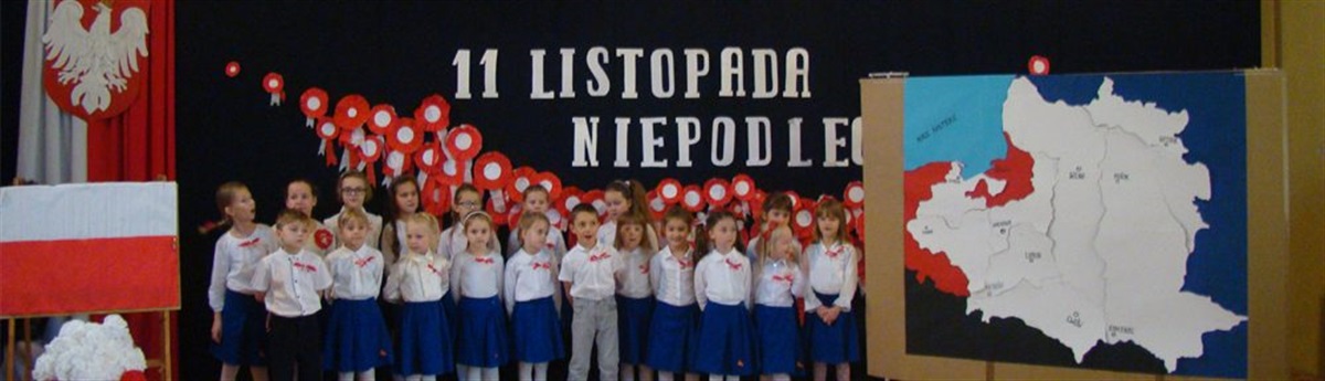 Kilkanaścioro ubranych na galowo dzieci stoi na sali gimnastycznej w dwóch rzędach. Za nimi na granatowej kurtynie widać napis 11 LISTOPADA NIEPODLEGŁA 
