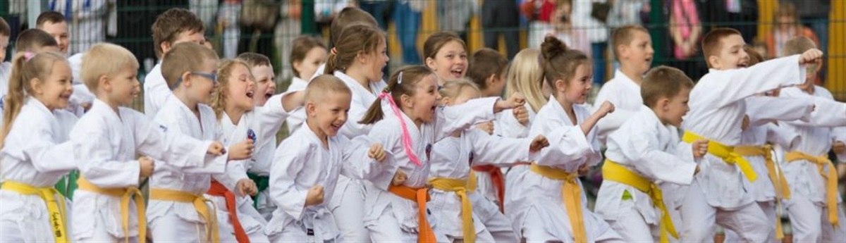 Grupa około 20 uśmiechniętych dzieci stoi na boisku szkolnym, dzieci ubrane są w białe kimona do karate