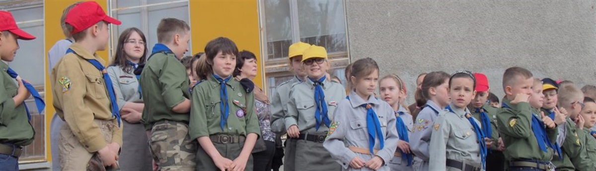 Grupa kilkunastu uczniów stoi przed budynkiem szkoły. Dzieci mają na sobie mundury zuchowe. 